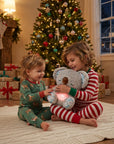 Two children in festive Christmas pajamas sit on a white rug in a cozy living room, hugging a glowing plush koala toy. A decorated Christmas tree, fireplace, stockings, and wrapped gifts are visible in the background.