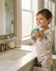 A toddler wearing animal-print pajamas stands at a bathroom counter holding a turquoise toothbrush holder with a toothbrush, with a mirror, sink, and window in the background.