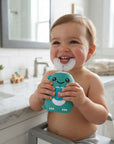 A smiling baby sitting in a bathroom high chair holds a teal cartoon-shaped silicone pacifier clip with a white bib or diaper cover attached. The background shows a bright, modern bathroom with white tiles, a marble countertop, and a sink.