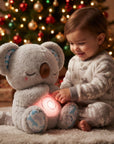 A toddler in light gray patterned pajamas sits on a rug next to a plush gray koala toy with a glowing pink belly, with a decorated Christmas tree in the background.
