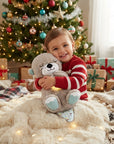 A smiling young child in a red and white striped sweater hugs a gray plush teddy bear with light blue accents, sitting on a fluffy cream blanket in front of a decorated Christmas tree with wrapped presents.