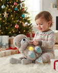 A toddler in patterned holiday pajamas sits on a white rug in a living room, holding and playing with a plush gray bear toy that has embroidered patches and a glowing light. A decorated Christmas tree, wrapped gifts, and a fireplace are visible in the background.