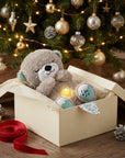 A plush teddy bear toy with a warm light glowing from within, nestled in a cream gift box on a wooden table. The background features a decorated Christmas tree with gold and silver ornaments, pinecones, and warm fairy lights, with a red ribbon in the foreground.