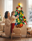A woman and child decorate a large felt Christmas tree with colorful holiday ornaments in a warm, decorated living room. The felt tree is green and features a snowman, duck, Santa, gifts, and warm string lights.