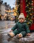 A child wearing a dark green Christmas-themed puffer suit with a beige knit beanie and tan boots sits on a snowy street in front of a red telephone booth decorated with lights and greenery.