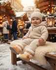 A baby wearing a cozy cream-colored knit snowsuit, matching beanie, and tan boots sits on a wooden bench at a snowy Christmas market with warm lights and decorations in the background.