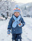 A smiling toddler standing in the snow wearing a blue winter outfit with a light blue scarf, blue hat, and matching gloves. The hat and gloves feature cartoon character graphics, and the scarf reads 'PAW PATRIOT'.