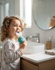 A young child with curly hair stands at a bathroom sink, smiling while brushing their teeth with a turquoise toothbrush holder in one hand. The scene includes a white sink, wooden cabinet, folded towel, and a round mirror.