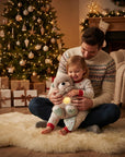 A father and child sitting together on a fluffy cream rug in a cozy living room, surrounded by a decorated Christmas tree, wrapped gifts, and a fireplace. The child is holding a plush teddy bear with a glowing yellow light.