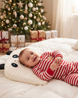 A baby wearing red and white striped Christmas pajamas lies on a white quilted bed next to a panda-shaped pillow, with a decorated Christmas tree and wrapped gifts in the background.