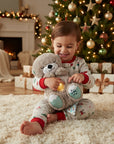A young child wearing festive holiday pajamas sits on a fluffy cream rug, holding a plush teddy bear with glowing light patches. The background features a beautifully decorated Christmas tree with warm lights, wrapped gifts, and a cozy fireplace.