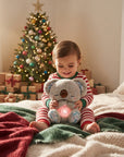 A toddler in red and white striped pajamas sits on a bed holding a gray koala plush toy with a glowing pink belly light, with a decorated Christmas tree and wrapped gifts in the background.