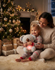 A woman and a young child sit on a fluffy cream rug in a cozy living room, hugging a gray plush koala toy that glows with a warm pink light. The background features a decorated Christmas tree, wrapped gifts, and a fireplace with warm lights.