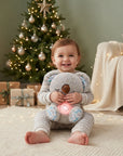 A smiling baby sits on a cream rug in a cozy room, holding a plush koala toy with a glowing pink belly light. A decorated Christmas tree, wrapped gifts, and a soft blanket are visible in the background.