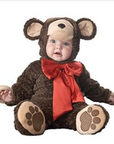 A baby wearing a plush brown teddy bear costume with a beige snout and ears, a red satin bow, and brown paw-print details on the feet, photographed against a plain white background.