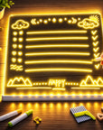 A glowing LED light-up writing board on a wooden desk, with a hand holding a white marker pointing at the illuminated text. The board displays yellow neon-style drawings and words including 'Rain' and 'HAPPY'.