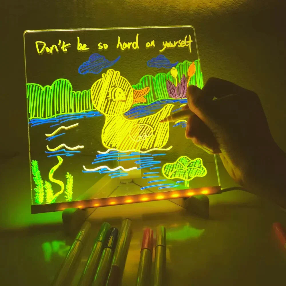 A glowing acrylic light box with a hand drawing on it, showing a yellow duck in a green and blue landscape with the text 'Don't be so hard on yourself' at the top. Several markers are lined up in the foreground on a dark surface.