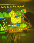 A glowing acrylic light box with a hand drawing on it, showing a yellow duck in a green and blue landscape with the text 'Don't be so hard on yourself' at the top. Several markers are lined up in the foreground on a dark surface.