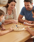 A family of four gathered around a wooden table playing with a wooden peg board toy. The mother, wearing a floral apron, and the father in a blue t-shirt are smiling and watching their children, who are actively engaged in the game.