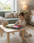 A toddler kneels on a light rug in a cozy bedroom/living area, playing with colorful fabric books on a small wooden table. The room features a green sofa, a bed, and warm ambient lighting.