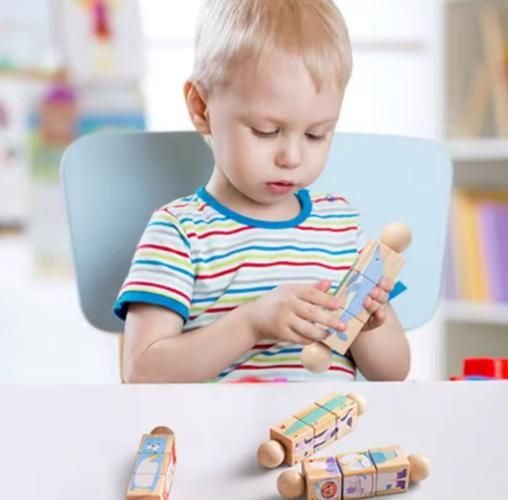 A young child sitting in a chair at a table, holding a wooden toy car. More wooden toy cars are scattered on the white table in front of the child.