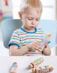A young child sitting in a chair at a table, holding a wooden toy car. More wooden toy cars are scattered on the white table in front of the child.
