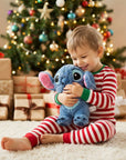 A young child wearing red and white striped pajamas sits on a white rug in front of a decorated Christmas tree, hugging a blue plush toy. Gift boxes are wrapped in beige paper with red and brown ribbons on the floor.