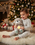 A toddler in light gray patterned pajamas sits on a white faux fur rug in a living room, hugging a plush teddy bear that glows with a warm yellow light. A decorated Christmas tree, fireplace, and wrapped presents are visible in the background.