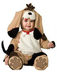 A baby wearing a plush dog costume, featuring a tan hood with long floppy dog ears, a white faux-fur chest panel, black paw pads, and a red collar, photographed against a plain white background.