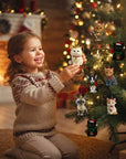 A young girl in a beige knit sweater kneels beside a decorated Christmas tree, smiling as she holds a white cat ornament. The tree is lit with warm fairy lights and decorated with several colorful cat ornaments, with a cozy fireplace in the background.