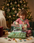 A child in red-and-white striped pajamas sits on a rug in front of a decorated Christmas tree, opening an advent calendar box with dinosaur toys scattered around. Presents and a cozy living-room setting are visible.