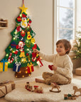 A young child sits on a white rug in a cozy room, touching a large green felt Christmas tree decoration with colorful hanging ornaments and warm string lights. The scene includes wrapped gifts, wooden toys, a window, and another lit Christmas tree in the background.