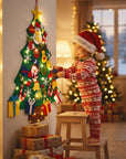 A child in a red and white holiday onesie and Santa hat stands on a small wooden stool, decorating a hanging Christmas tree with colorful ornaments in a warmly lit living room. Wrapped gifts sit below the tree, and blurred Christmas lights and another tree appear in the background.