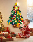 A toddler in red and white striped pajamas sits on a carpeted floor next to a decorated Christmas tree, holding a small yellow ornament. A felt Christmas tree with colorful holiday decorations and warm string lights hangs on the wall behind the child.