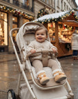 A baby wearing a cream cable-knit sweater and matching pants sits in a beige stroller on a snowy Christmas market street, with decorated storefronts and market stalls in the background.