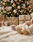 A close-up of a child's legs wearing beige knit socks and tan slippers with white faux-fur trim and pom-poms, posed on a fluffy cream rug in front of a decorated Christmas tree and wrapped gift boxes.