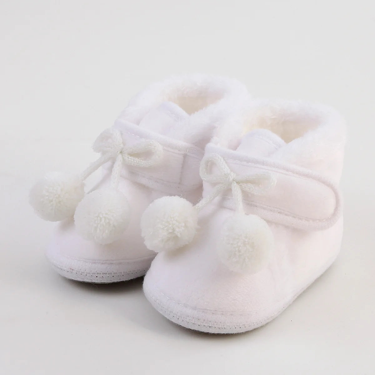 Pair of white baby shoes with white fur lining and pom-pom details on a light background