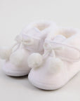 Pair of white baby shoes with white fur lining and pom-pom details on a light background